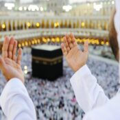 A man praying Dua standing in Masjid Al-Haram mecca, with Kaaba in background.